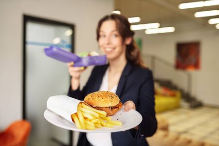 French fries and burger. Happy girl holding purple open lunchbox in one hand, the other hand holding forward plate with burger and french friesの写真素材