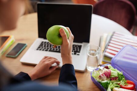 Appetizing apple. Green juicy apple in female hands over a table with a laptop and a lunchbox with vegetables.の写真素材