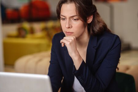 Work moment. Serious long-haired girl in a blue blazer sitting in front of a laptop, an attentive concentrated hand near her chin.の写真素材
