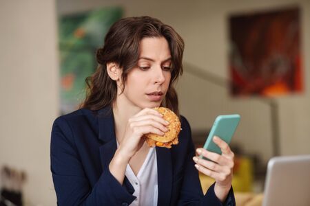 Interesting. Serious interested young woman with a burger in one hand in the other with a smartphone, carefully looking at the screenの写真素材