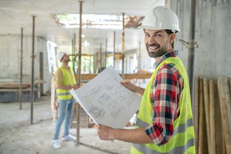 Building site. Building worker in yellow vest turning his head to camera, holding blueprint, his...