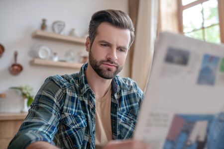 Important news. Man in a checkered shirt sitting at the table, reading newspaper and looking seriousの写真素材