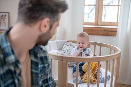 Father and son. Dark-haired father watching his little son playing in the playpen