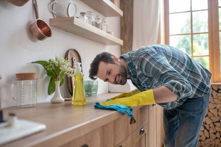 In the kitchen. Dark-haired man in a checkered shirt and in yellow gloves cleaning the table in the...