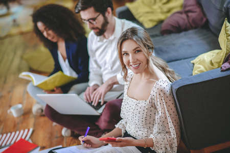 Work with information.. Smiling long-haired girl with smartphone recording pen in papers and two...