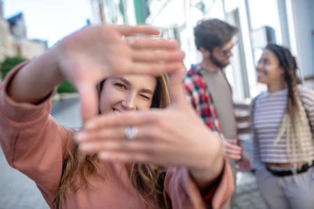 Beautiful view. Cheerful young adult girl making aerial lens with her hands looking close one eye, friends at a distanceの写真素材