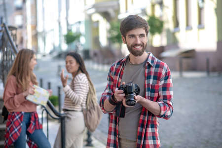 Pleasant impression. Young adult bearded guy with camera standing on the street, looking smiling forward, two girlfriends behindの写真素材