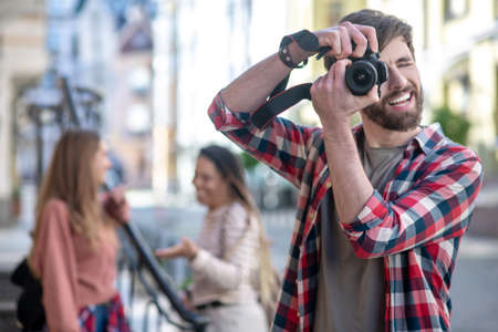 Favourite hobby. Guy in a plaid shirt photographing on a digital camera on a city street, two girls standing behindの写真素材