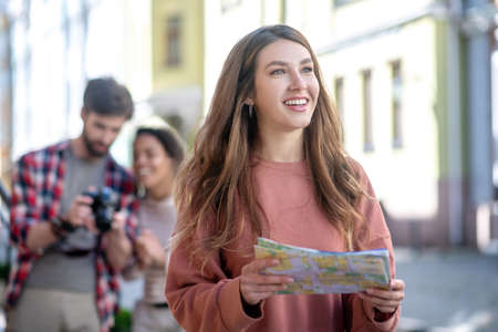 Girl with map. Young long-haired girl shining with delight, with map in her hands, standing on city street, friends at distanceの写真素材