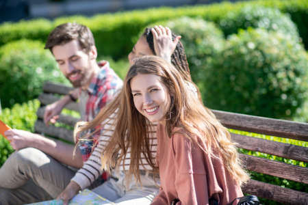 Friends on bench. Three young friends sitting on bench on sunny warm day, chatting vacationers in good mood.の写真素材