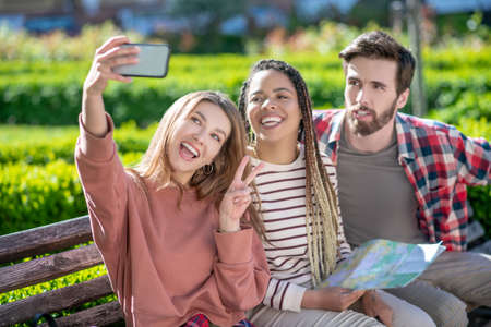 Selfies. Girl with smartphone taking selfie with mulatto girlfriend with dreadlocks and bearded guy...