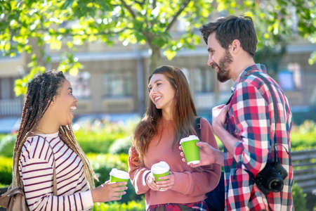 Delight, communication. Enthusiastic mulatto girl with dreadlocks with open mouth and opposite girlfriend and boyfriend all standing with coffeeの写真素材