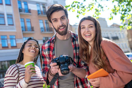 Happy young friends. Guy with camera, mulatto girl with coffee and girlfriend with tablet, standing close satisfied smilingの写真素材