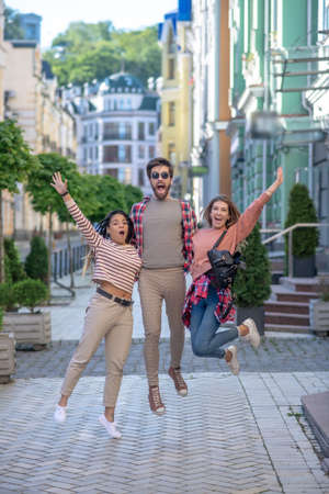 Freedom. Bearded guy in sun-protection glasses and two girls bouncing up together, raising his hands, opening his mouth, enthusiastic on street.の写真素材