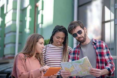 Search, map. Guy in sunglasses with a map and two girls with a tablet standing together in the...