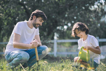 Plants. Dark-haired boy watering the plant and smiling, his father watching himの写真素材