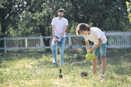 In the garden. Dark-haired boy watering the plant, his father standing nearby