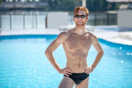 Beautiful sunny day. Red-haired smiling man in sunglasses and swimming suit standing near pool with...