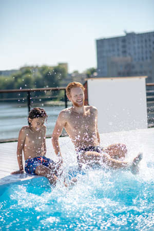 Refreshing spray. Young dad with his son sitting on the poolside making splashes with his feet, funny laughingの写真素材