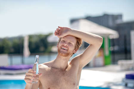 Summer, heat. Red-haired bearded young man with bottle of water holding hand near forehead standing near pool on hot summer dayの写真素材