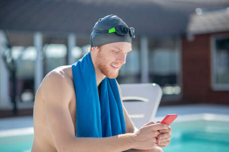 Interesting here. Young bearded smiling swimmer in swimming cap and towel sitting on sunbed with smartphone by poolsideの写真素材
