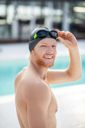 Swimming. Contented joyful man in black swimming cap and black swimming goggles near swimming pool