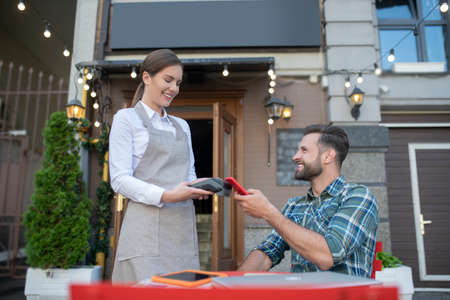 Paying bill. Bearded male paying bill with smartphone, waitress holding payment terminal, smiling