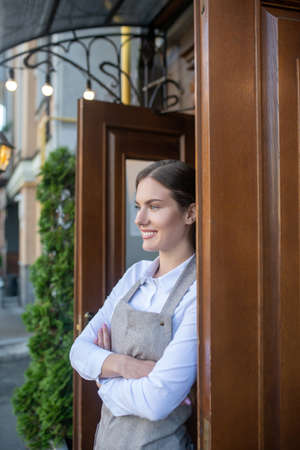Cosy cafe. Smiling cute waitress in grey apron leaning on door with her arms crossedの写真素材