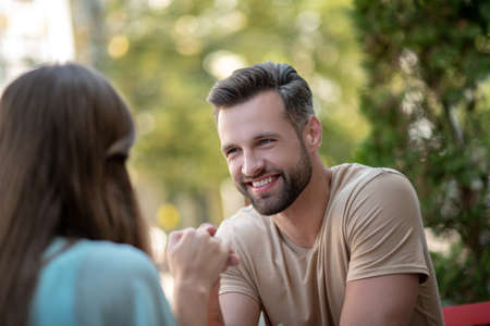Loving relationship. Smiling bearded male sitting opposite female in blue dress, holding her handの写真素材