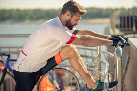 Warming up muscles. Concentrated young bearded man in sports gloves holding handrail raised leg and...