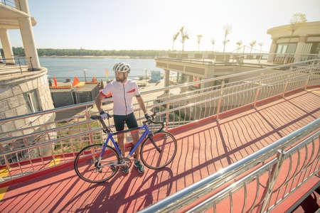 Walk, beautiful view. Young adult cyclist in sportswear standing with bike on the bridge looking at beautiful viewの写真素材