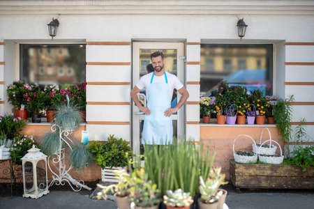 Flower shop. Young bearded male in apron standing at the door of flower shop with hands on his hipsの写真素材
