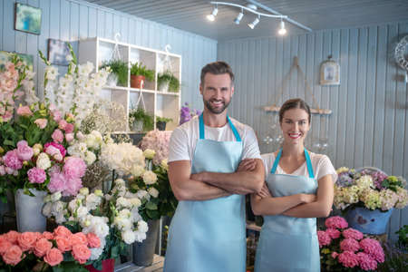 Flower shop. Bearded male and brown-haired female wearing aprons, standing intside flower shop with crossed armsの写真素材