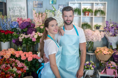 Flower shop. Bearded male and brown-haired female in aprons embracing each other, woman putting her hand on mans shoulderの写真素材