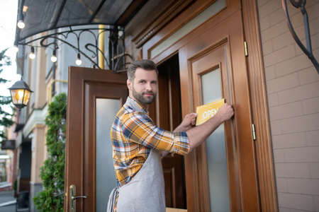 Restaurant business. Young bearded waiter in grey apron hanging open sign on the door, smiling