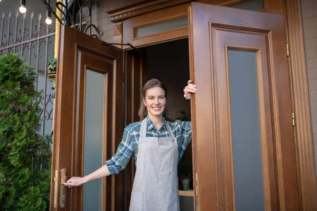 Restaurant business. Smiling young waitress holding doors, welcoming clients