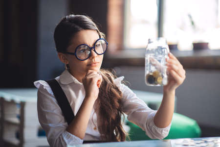 In thoughts. Dark-haired girl holding jar with coins and thinkingの写真素材