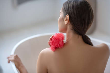 In the bath. Woman holding a red sponge and sitting in the bathの写真素材