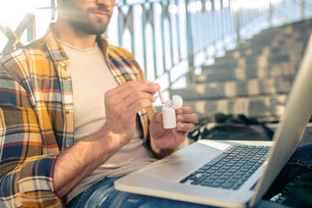 Useful gadget. Bearded man with laptop sitting on stairs holding wireless white headphones and caseの写真素材