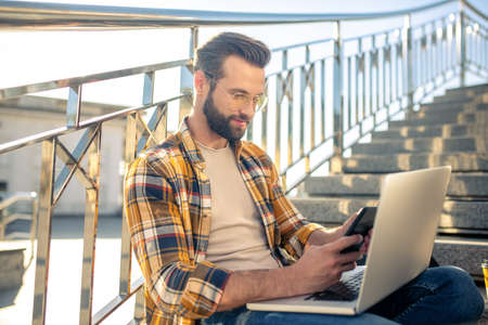 Interesting information. Young bearded man in glasses with smartphone in hands sitting on steps of...