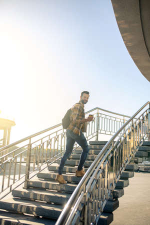 Rising up. Joyful dark-haired man with backpack and coffee climbing up the city stairs on sunny...
