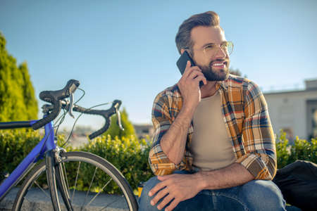 Nice talking. Calm smiling bearded man with smartphone near ear sitting on street near bike