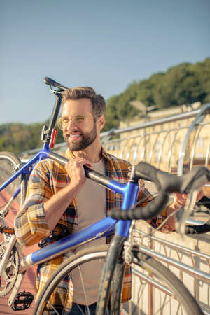 Active walk. Joyful young adult bearded dark haired man standing outdoors holding bike on shoulderの写真素材