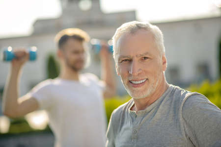 Morning workout. Grey-haired male smiling, young bearded male holding blue dumbbells behind himの写真素材