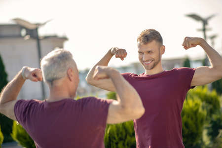 Exercising outside. Bearded young male and grey-haired man standing opposite each other, showing...