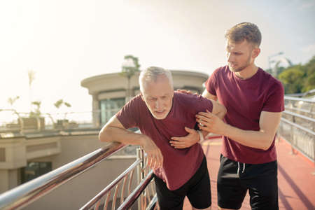Heart attack. Mature grey-haired male holding hand on his chest, young male supporting him