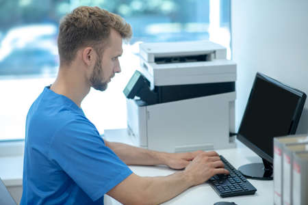 Health care, information. Young adult serious man in profile in blue medical uniform sitting at...