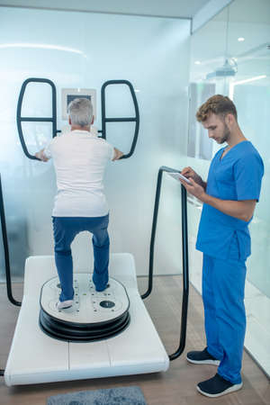 Simulator session. Gray-haired man near simulator with his back to camera and young instructor in blue uniform looking at tabletの写真素材