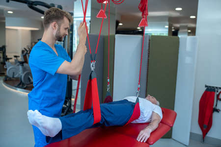 Rehabilitation procedure. Cheerful male rehabilitator lifting up on the leg straps of an elderly patient lying calmly.の写真素材
