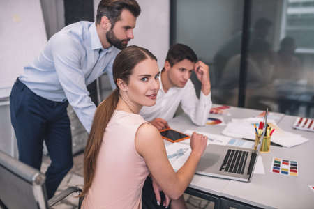 Fashion studio. Brown-haired female looking into camera, her male colleagues checking something on laptopの写真素材